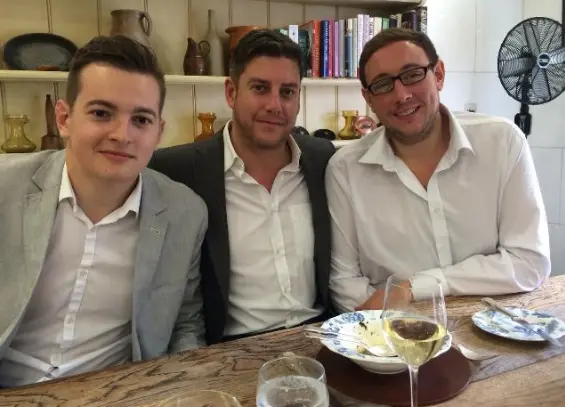 : Three men sitting together at a restaurant table, smiling at the camera, with plates and drinks in